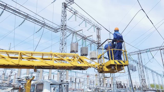 workers in crane cradle on jib against wires web on distribution station