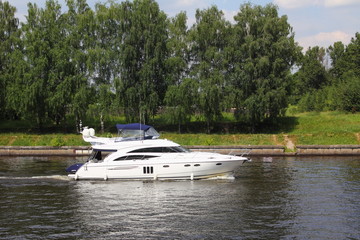 Fototapeta na wymiar Travel, luxury water recreation on boat - a white 58-ft motor yacht slowly sails along the concrete shores of the navigable canal in summer amid the green trees on the shore, side view