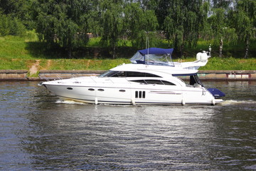 Travel, luxury water recreation on boat - a white 58-ft motor yacht slowly sails along the concrete shores of the navigable canal in summer amid the green trees on the shore, side view in the day