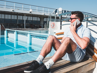 Smiling man talking on the phone on an open deck of a cruise ship on a background of the pool and...