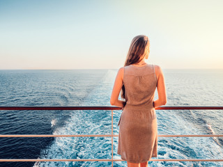Stylish, young woman on an empty deck of a cruise ship against a background of sea waves, blue sky and sunset - obrazy, fototapety, plakaty