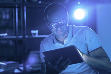 At night. Nice positive cheerful man holding a tablet and using it while being awake late at night