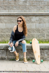 Young cheerful woman in fashion sunglasses and rock black style leather jacket with longboard skateboard and white music headphones. Hipster urban style girl. Lifestyle outdoor city portrait. - 214426311