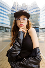Young woman in fashion glasses, rock black style leather jacket and hipster cap hat in front of modern business building from glass look to camera. Urban style girl. Lifestyle outdoor city portrait.