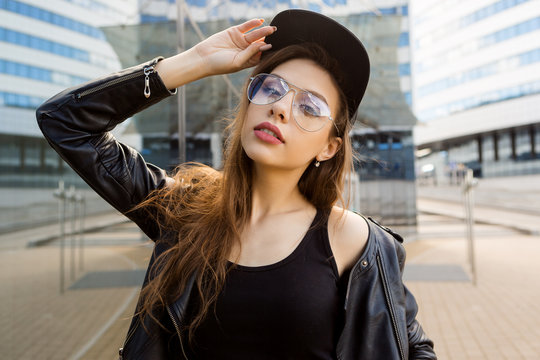 Beautiful lady in fashion glasses rock black style leather jacket and hipster cap hat in front of modern business building from glass look to camera. Urban style girl. Lifestyle outdoor city portrait.