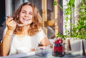 Young beautiful happy smiling woman eating yogurt and fresh berries at breakfast at home