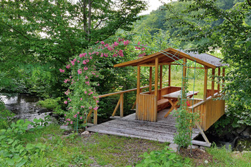 Fototapeta na wymiar Wooden gazebo with a roof and a table standing on the boards lying over the stream below. And growing flowers on an arch of wire made nearby.