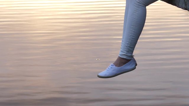 Woman legs in white shoes dangle over calm water surface