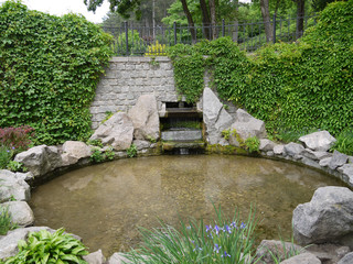 A round, small pond surrounded by stones, with a small waterfall