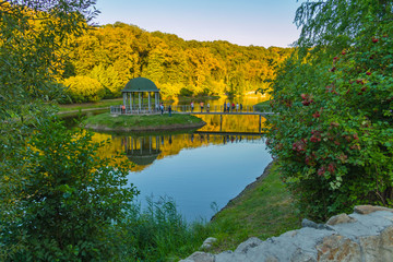 Arbor for recreation located on a green island in the middle of the lake with a bridge leading to it. With tourists admiring the beauty of the setting sun illuminating the tall trees on the opposite