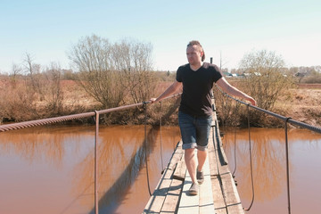 Naklejka premium Young man is walking on a suspended wooden bridge over the river.