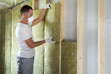 Worker in protective goggles and respirator insulating rock wool insulation in wooden frame for...
