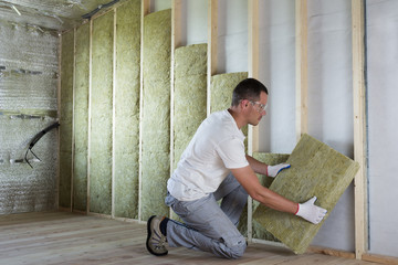 Worker in protective goggles and respirator insulating rock wool insulation in wooden frame for...