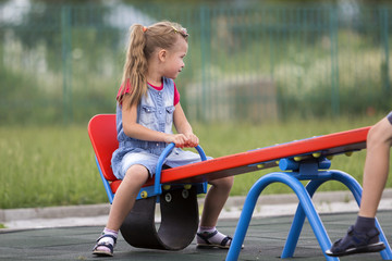 Small cute young blond child girl sits on see- saw swing on warm sunny day on bright green blurred...