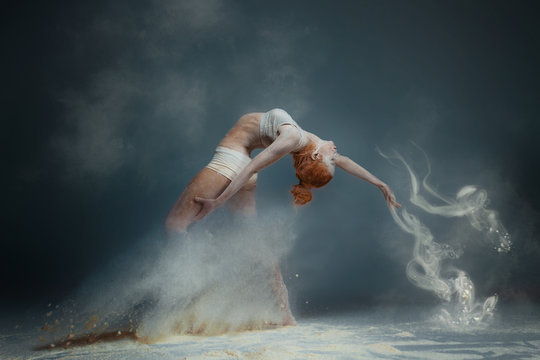 Dancing in flour concept. Redhead beauty female girl adult woman dancer in dust / fog. Girl wearing white top and shorts making dance element in flour cloud on isolated grey black background