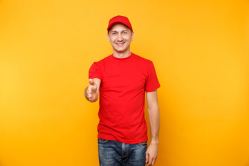 Delivery man in red uniform isolated on yellow orange background. Smiling male employee in cap, t-shirt working as courier dealer standing with outstretched hand for greeting gesture. Service concept.