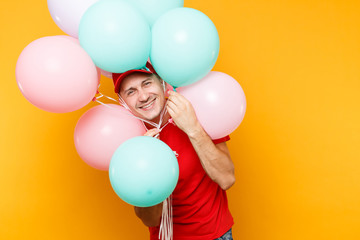 Obraz premium Delivery man in red uniform isolated on yellow orange background. Professional male employee in cap, t-shirt courier dealer holding bunch of colorful pastel air balloons. Service concept. Copy space.