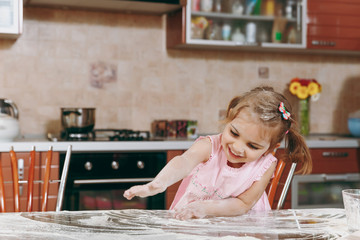 Obraz premium Little pretty kid girl in pink dress playing, smearing hands and drawing with flour in light kitchen at table. Child daughter cooking food, cookies in weekend morning at home. Happy childhood concept.