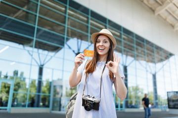 Young shocked traveler tourist woman with retro vintage photo camera, showing OK sign, holding credit card at international airport. Passenger traveling abroad on weekends getaway. Air flight concept.