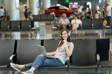 Young pensive traveler tourist woman with headphones listening music working on laptop, wait in lobby hall at international airport. Passenger traveling abroad on weekends getaway. Air flight concept.