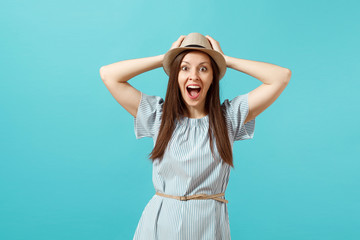 Obraz na płótnie Canvas Portrait of excited smiling young elegant woman wearing dress, straw summer hat put hands on head, copy space isolated on blue background. People sincere emotions, lifestyle concept. Advertising area.