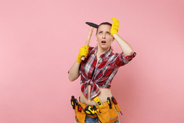 Strong young handyman woman wearing plaid shirt, denim shorts, kit tools belt full of different instruments hammer isolated on pink background. Female in male work. Renovation and occupation concept.