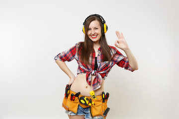 Handyman woman in shirt, denim shorts, noise insulated headphones, kit tools belt full of instruments showing OK gesture sign isolated on white background. Female in male work. Renovation concept.