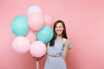 Portrait of attractive young happy woman wearing blue dress holding colorful air balloons showing thumb up isolated on bright pink background. Birthday holiday party, people sincere emotions concept.