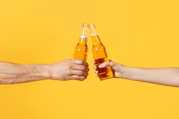 Close up cropped of woman and man two hands horizontal holding lager beer glass bottles and clinking isolated on yellow background. Sport fans cheer up. Friends leisure lifestyle concept. Copy space. - obrazy, fototapety, plakaty