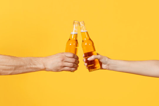 Close up cropped of woman and man two hands horizontal holding lager beer glass bottles and clinking isolated on yellow background. Sport fans cheer up. Friends leisure lifestyle concept. Copy space.
