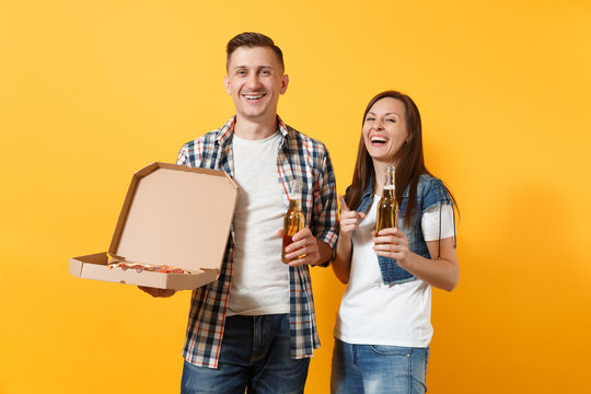 Young laughing couple woman man sport fans cheer up support team holding beer bottles italian pizza in cardboard flatbox pointing finger isolated on yellow background. Sport family leisure lifestyle.