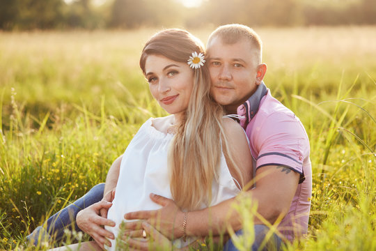 Affectionate young couple in love wait for child, sit and embrace together on green grass, have positive expression, want to become parents soon. Maternity, fatherhood and pregnancy concept.