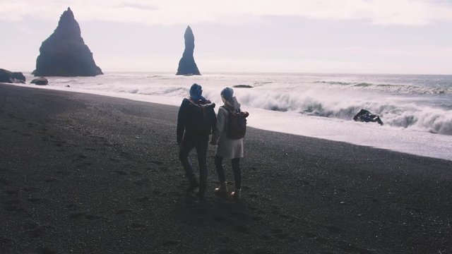 Young couple holding hands walking down black sand beach in Iceland, slow motion