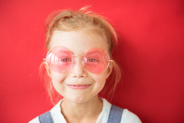 Smiling little girl in big round pink glasses on red background