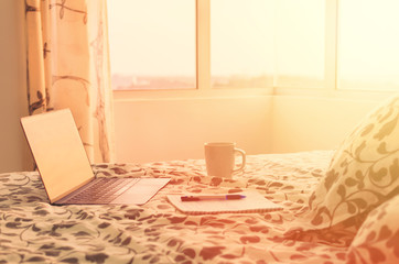 Sunny morning in modern apartment -  open laptop on bed opposite window,  cup of coffee and notebook. Beginning of the day.  Working at home, productive day, planning. Photo toned in warm colors
