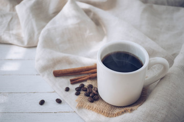 Hot coffee in white mug with coffee beans and cinnamon on wood background