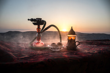 Hookah hot coals on shisha bowl making clouds of steam at desert outdoor. Oriental ornament on the carpet eastern tea ceremony. Stylish oriental shisha on sunset background.