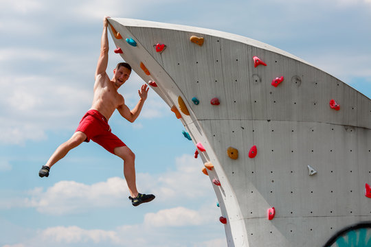 Photo of young sporty guy in red shorts hanging on wall for rock climbing against blue sky with clouds