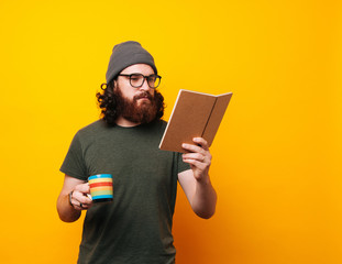 Bearded hipster man in green t-shirt wearing eyeglasses and hat and reading a book and holding cup...