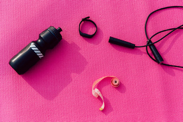 Top view of bottle, watches, skipping rope and measuring tape. Pink background. Sport equipment.