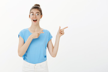 Portrait of good-looking happy young girl in glasses and bun, smiling broadly and pointing right with finger gun gestures, discussing great new place in town, suggesting friends to visit it