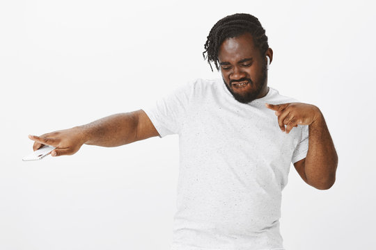 Indoor shot of energetic happy african-american plump man, dancing, stretching hand left while listening music in wireless earphones and holding smartphone, enjoying favorite songs over white wall