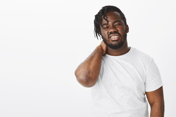 Tired displeased attractive dark-skinned guy in white t-shirt, rubbing neck with closed eyes and clenched teeth, feeling exhausted after long day working or sitting near pc, feeling pain in muscles