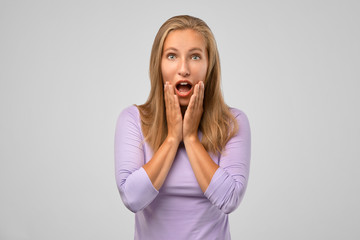 Woman is shocked, looking with disbelief. Portrait of cute caucasian blonde standing partially crossing body with hand, holding palm on cheek and dropping jaw from stunning news, condemning friend