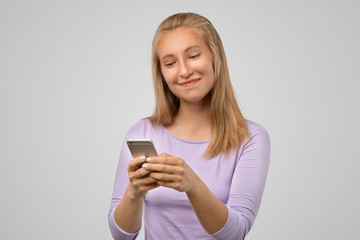 Fototapeta premium Happy loving girlfriend texting with satisfied expression. Portrait of pleased playful young girl in casual outfit, holding smartphone and smiling broadly, gazing at screen with satisfaction