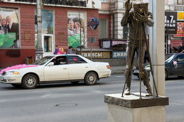 Monument to the photographer against the background of wedding cars on a city street. Sight is located in the center of Krasnoyarsk, Krasnoyarsk Territory, Russia.