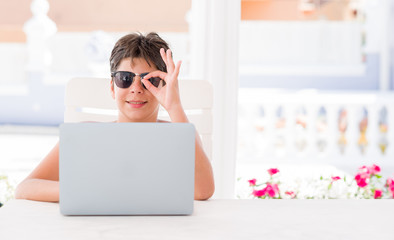 Young child on holidays using computer laptop with happy face smiling doing ok sign with hand on eye looking through fingers