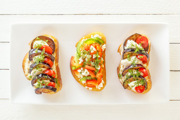 Bruschetta with fresh tomatoes and cheese, eggplant and mozzarella, sweet pepper and goat cheese on a white plate. Top view
