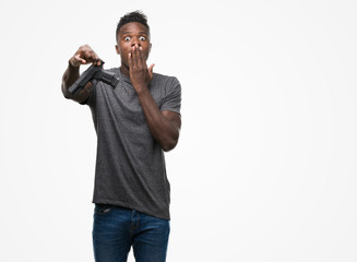 Young african american man holding a gun cover mouth with hand shocked with shame for mistake, expression of fear, scared in silence, secret concept