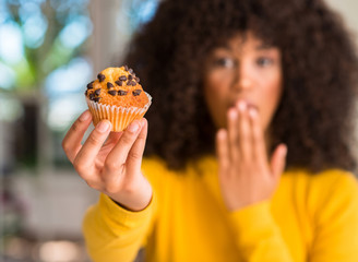 African american woman holding chocolate muffin cover mouth with hand shocked with shame for...